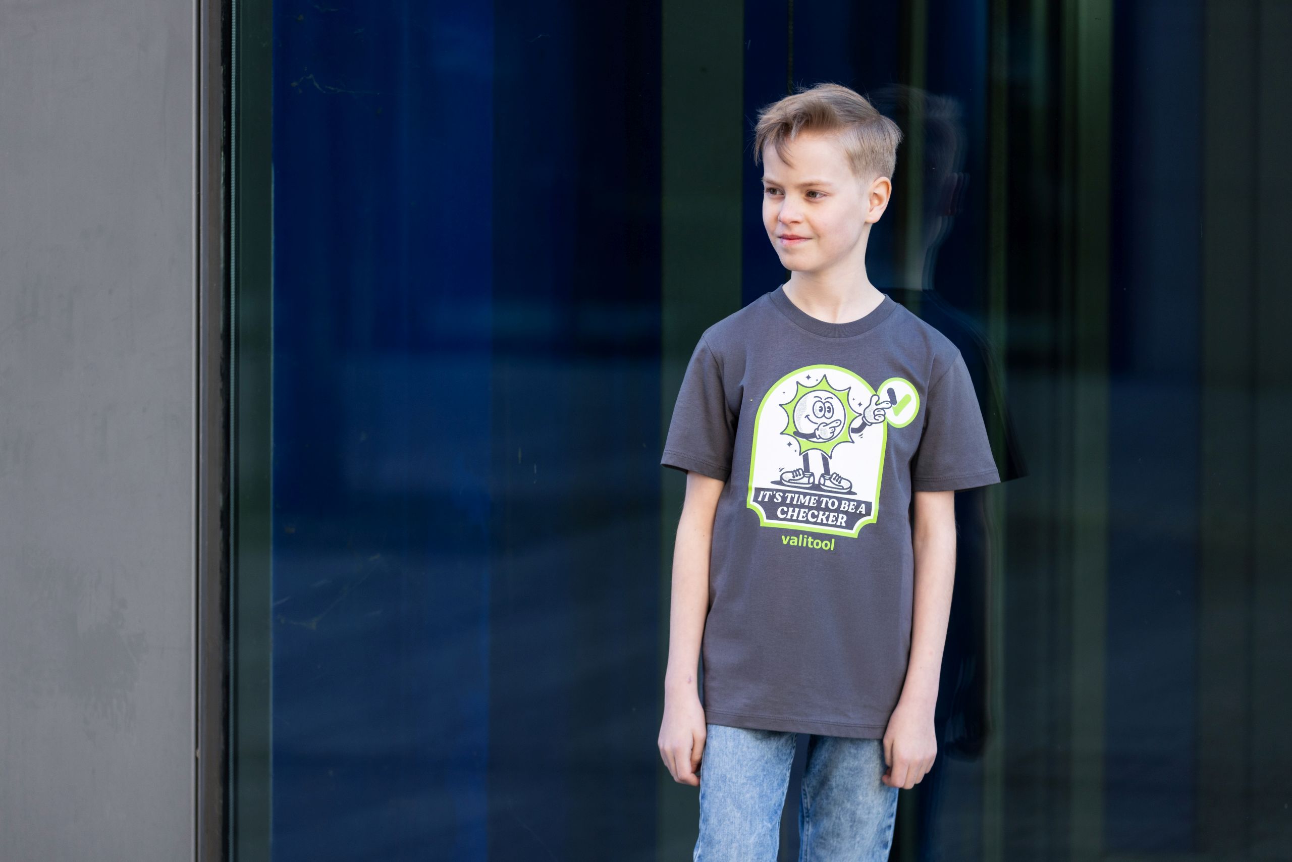 Junge trägt Valitool Kinder Unisex T-Shirt in Anthrazit mit Cartoon-Print ‚It’s Time to Be a Checker‘, Outdoor-Aufnahme vor Glasfassade