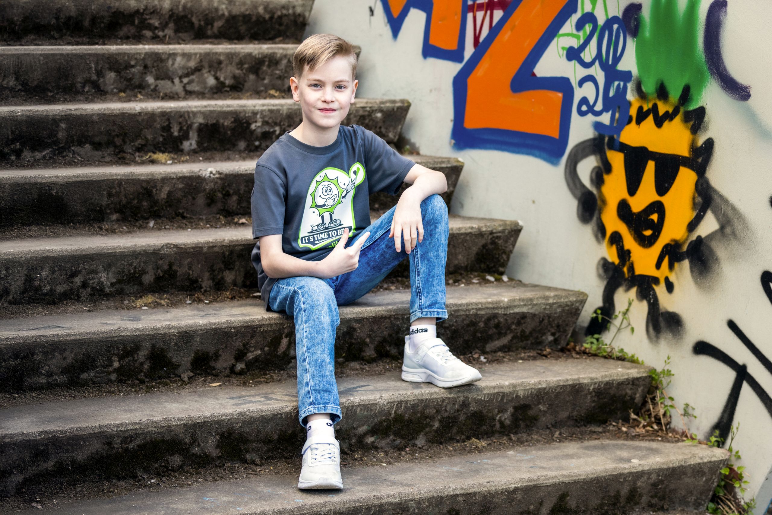 Junge trägt Valitool Kinder Unisex T-Shirt in Anthrazit mit Cartoon-Print ‚It’s Time to Be a Checker‘, sitzend auf Treppe mit Graffiti-Hintergrund